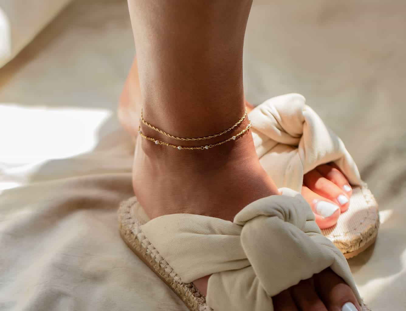 Two gold anklets, one beaded, stacked on a model's ankle above a sandal