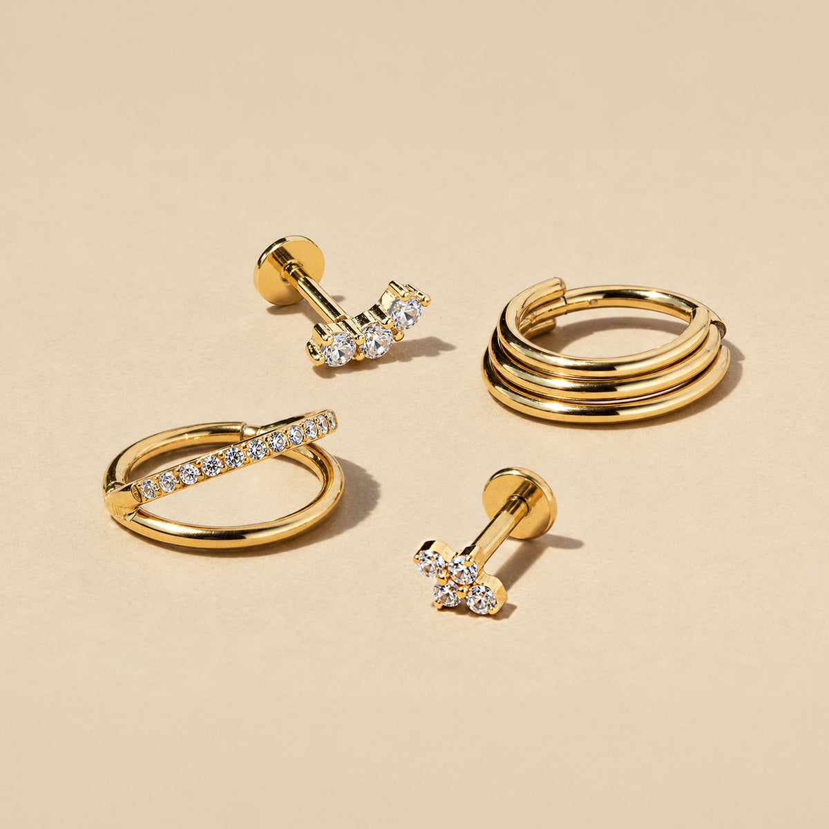 Four gold pieces on a beige surface: two crystal flat back studs, a crystal-lined huggie and a triple-band hoop