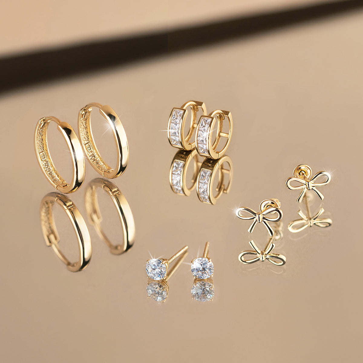 Four gold earring pairs on a reflective beige surface: plain huggies, crystal-set huggies, bow studs and round crystal studs