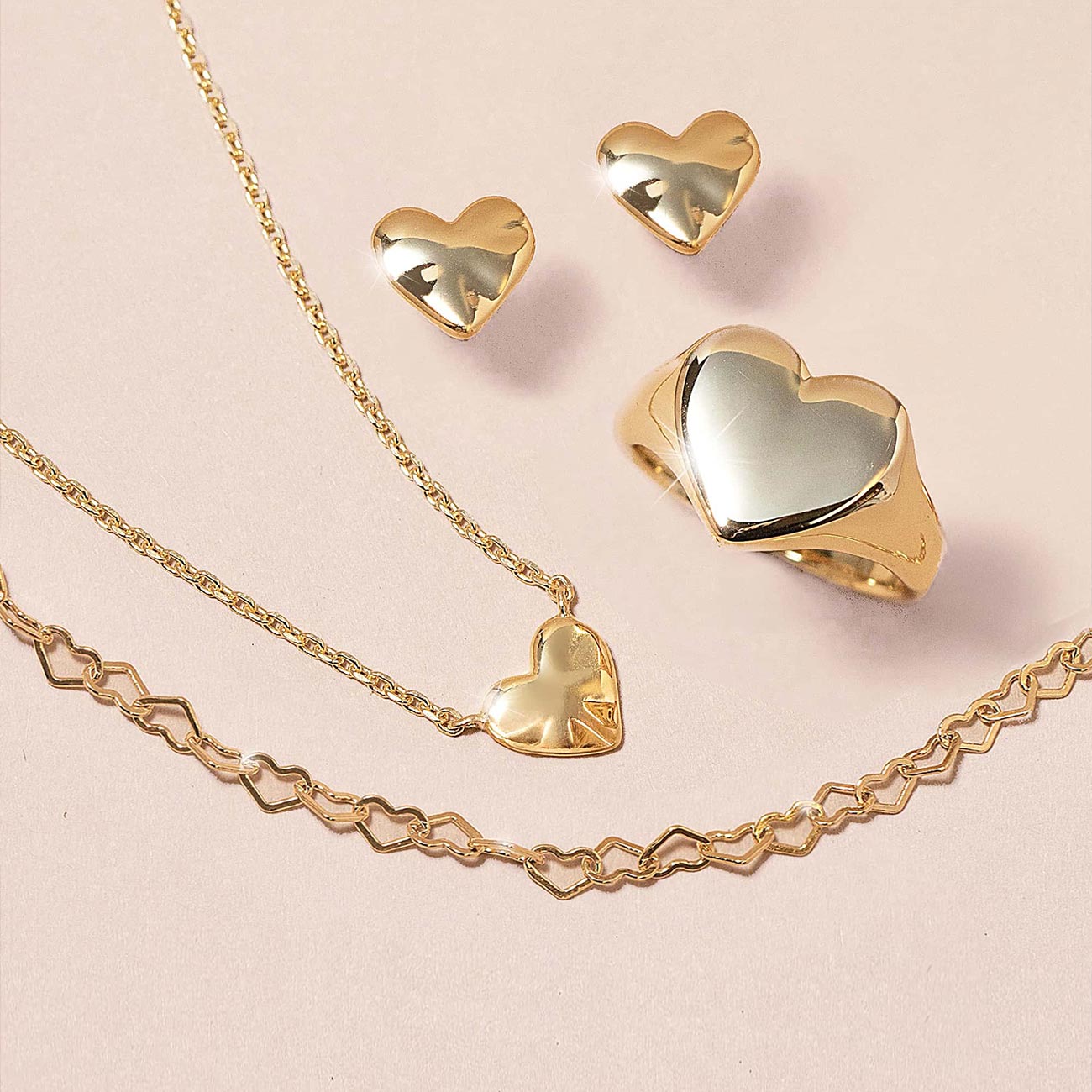 Gold heart-shaped jewelry including necklace, earrings, and ring on a light pink background. Heart Jewelry perfect for gifitng.