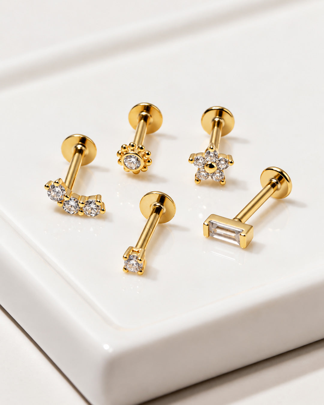 Five gold flat back studs with crystals in curved, beaded, star, solitaire and baguette shapes on a white tray