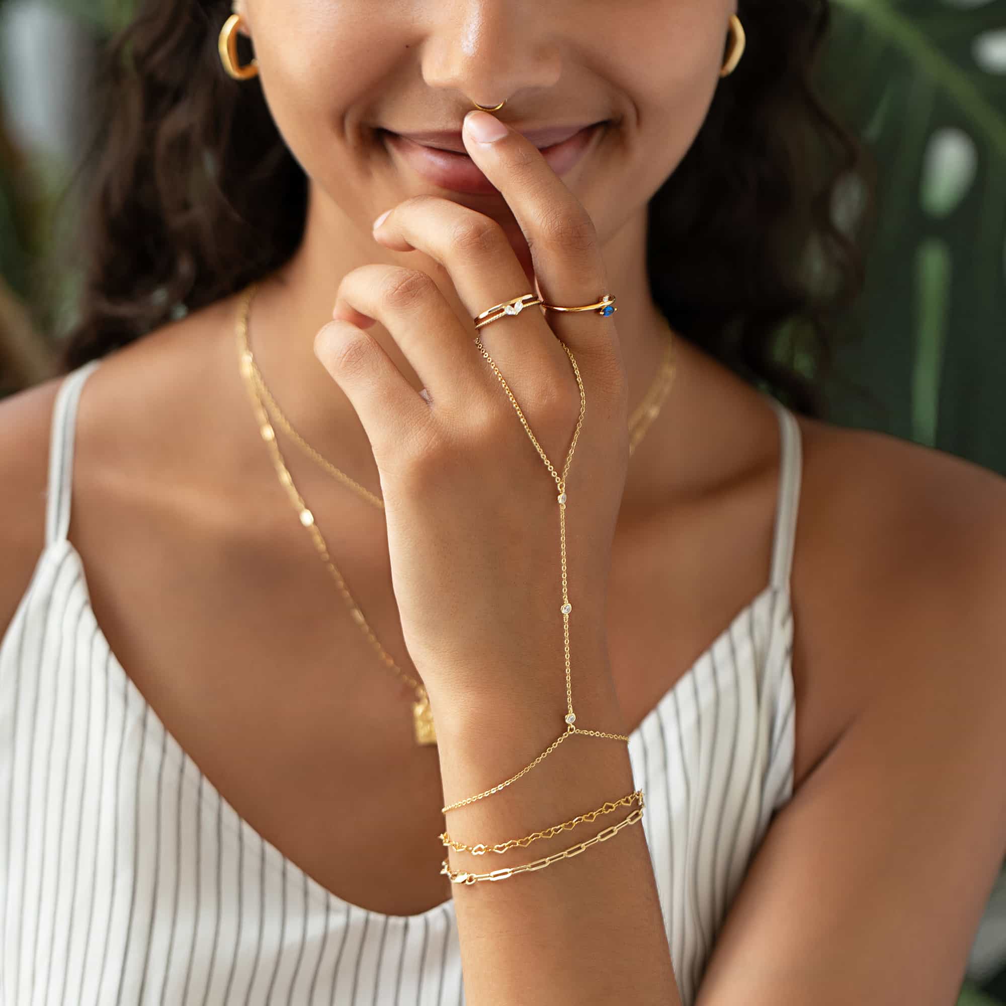 Woman wearing gold dainty crystal handchain, gold heart link chain bracelet, gold chain link bracelet, gold thin band and rope ring set, gold tiny pear heart ring, and gold marquise band ring