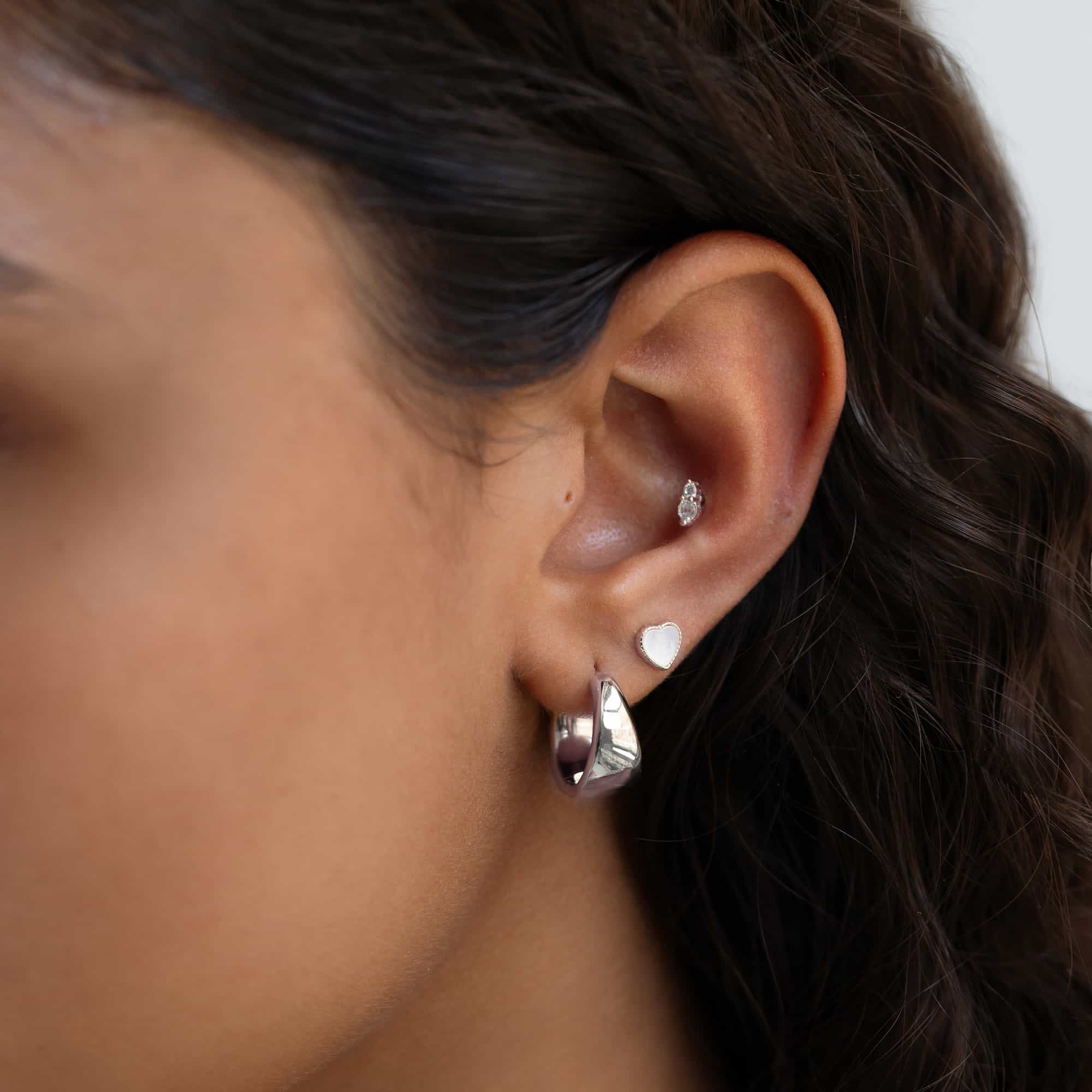 Silver ear stack on a model's lobe with a chunky huggie hoop, mother-of-pearl heart stud and tiny duo stud