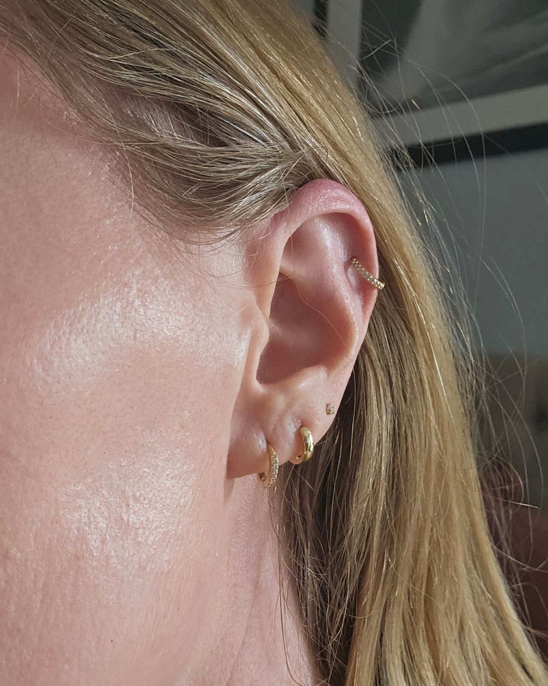 Close-up of an ear wearing two gold lobe huggie hoops, a small stone stud, and a gold helix hoop