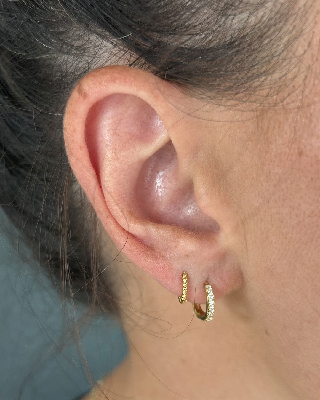 Two gold huggie hoops worn in a lobe, one beaded and one set with small crystals, on a close-up ear