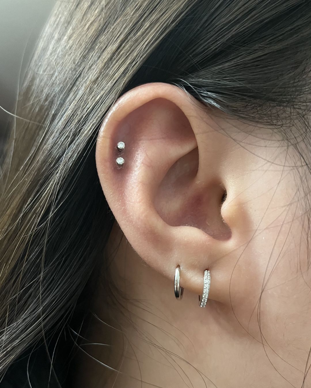 Close-up of an ear with two small crystal studs in the upper helix and a plain and pavé huggie hoop in the lobe