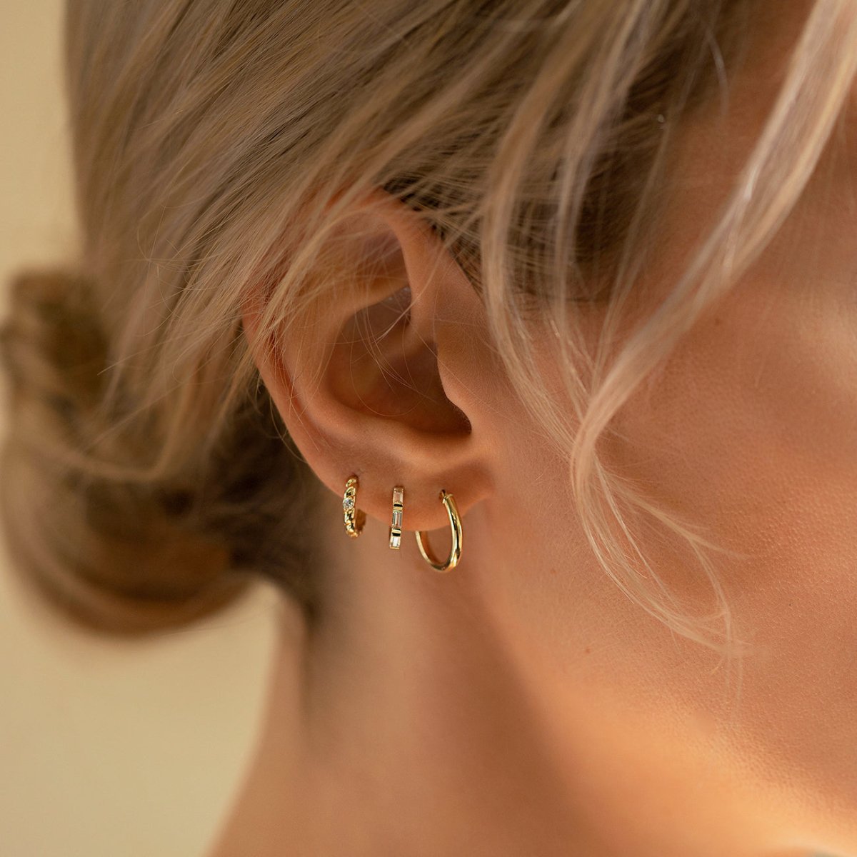 Close-up of woman's ear featuring 14K Solid Gold Tri Baguette Huggie Hoops Moissanite Crystals, 14K Midi Hoop Earrings, and 14K Pave Rope Huggies