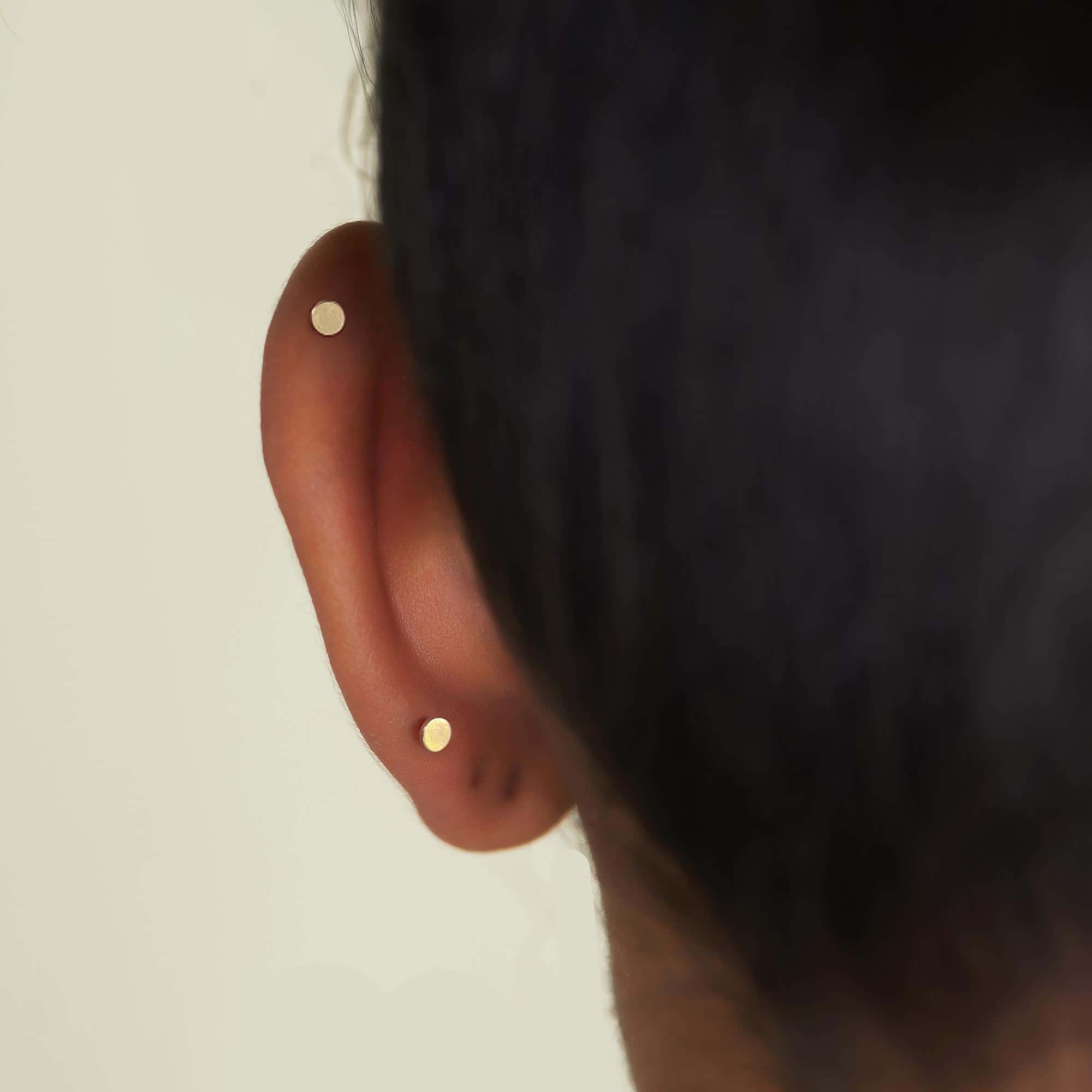 Two plain gold flat dot studs shown in a model's helix and lobe piercings, side view