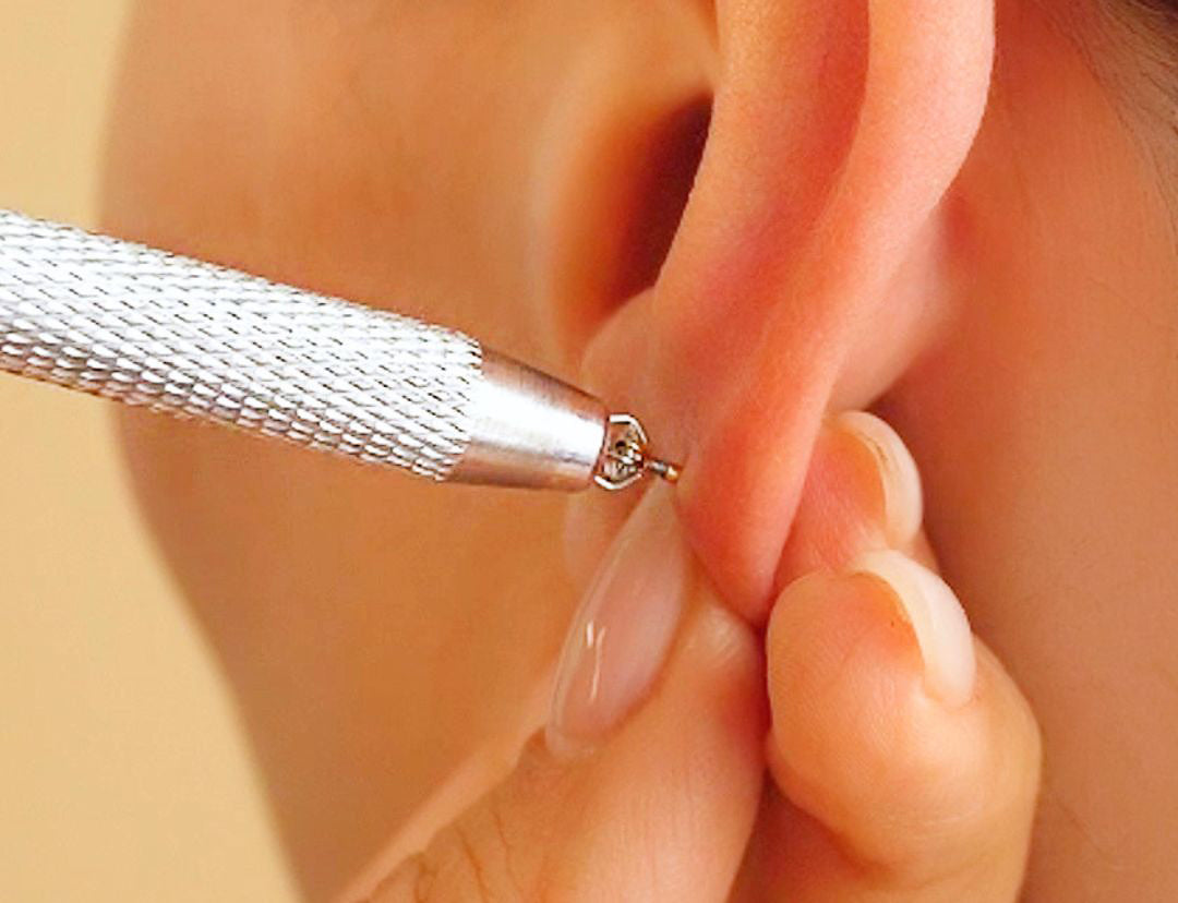 Close-up of an ear as a metal insertion tool holds a small stud, fingers steadying the lobe