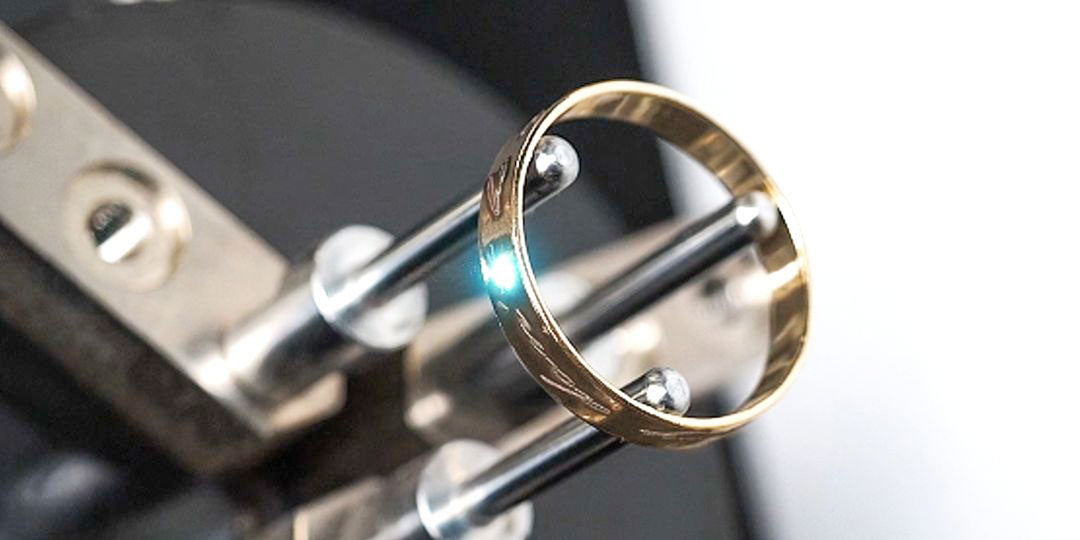 Plain gold band held in a metal engraving clamp, with a small bright light marking the ring's surface