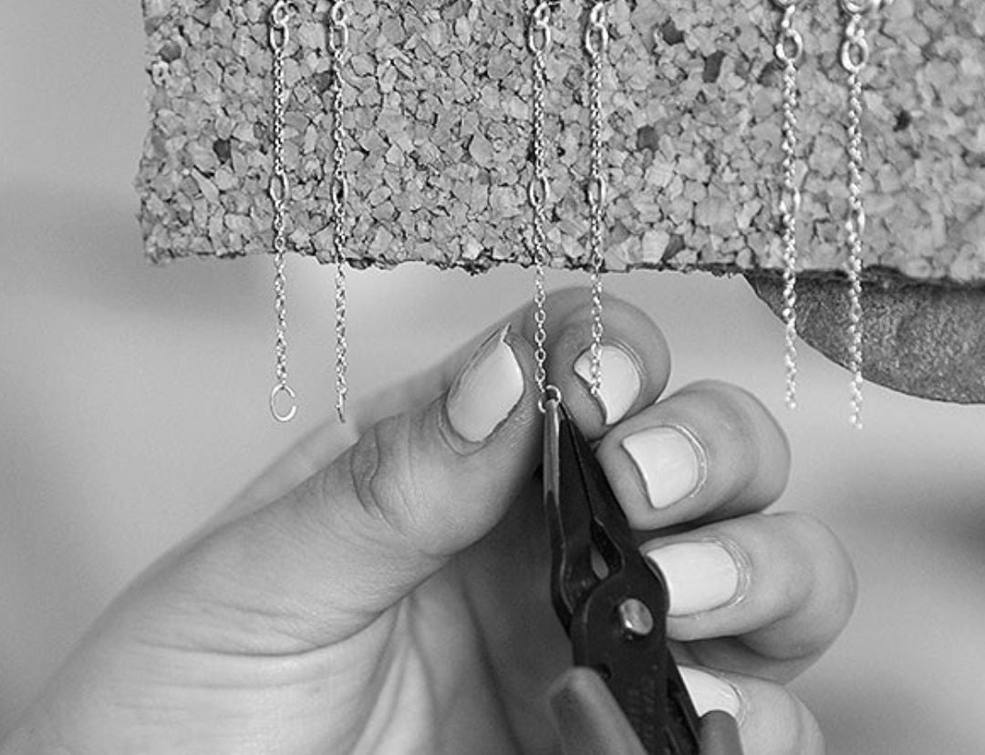 Black and white photo of two hands using pliers on fine chains hanging from a cork board