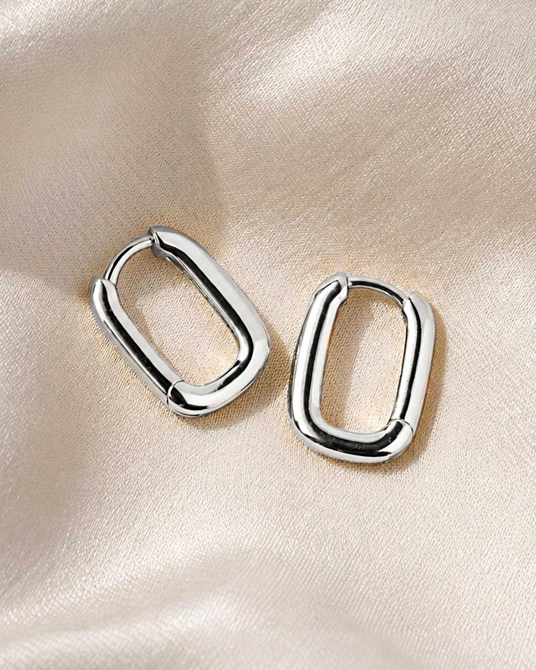 Pair of chunky silver oval huggie hoop earrings resting on cream satin fabric