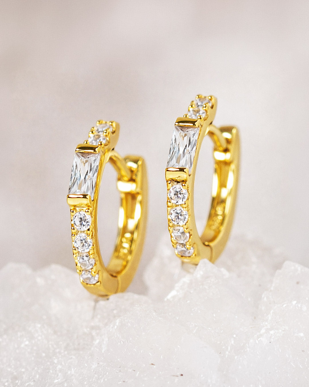 Gold baguette huggie earrings with white zirconia gemstones on a textured white surface from AMYO.