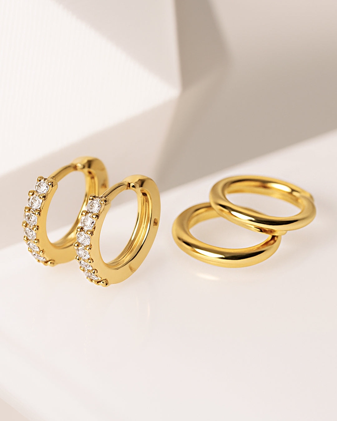 Two pairs of gold huggie hoops on a pale surface, one set with rows of clear crystals and one plain polished