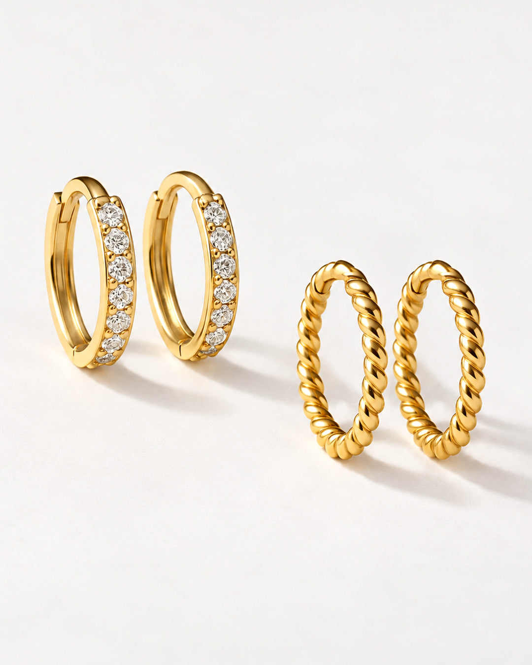Two pairs of gold huggie hoops on a pale surface, one set lined with clear stones, the other twisted rope