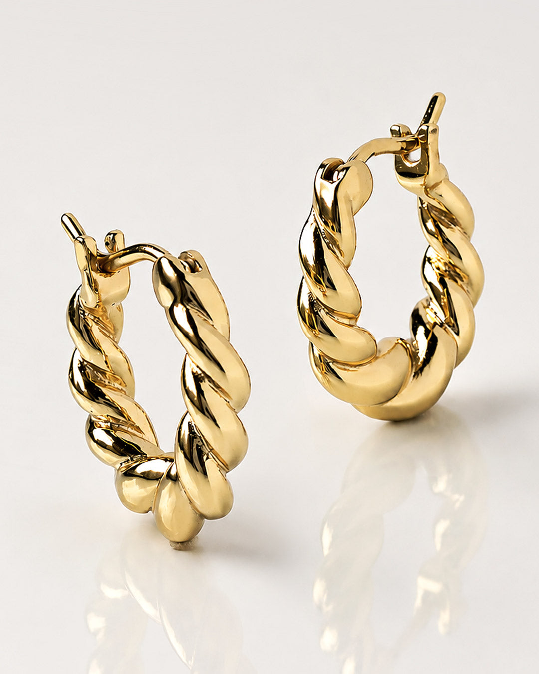Pair of gold twisted rope hoop earrings with hinged closures, standing upright on a reflective white surface