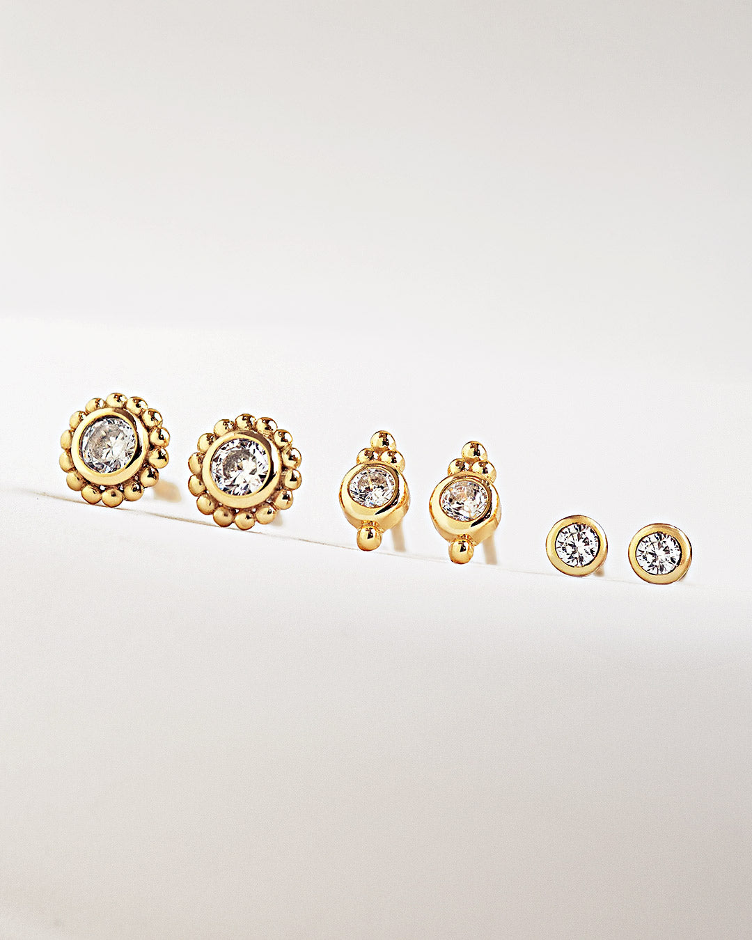 Sets of tiny gold stud earrings with white zieconia gemstones on a light gray background from AMYO