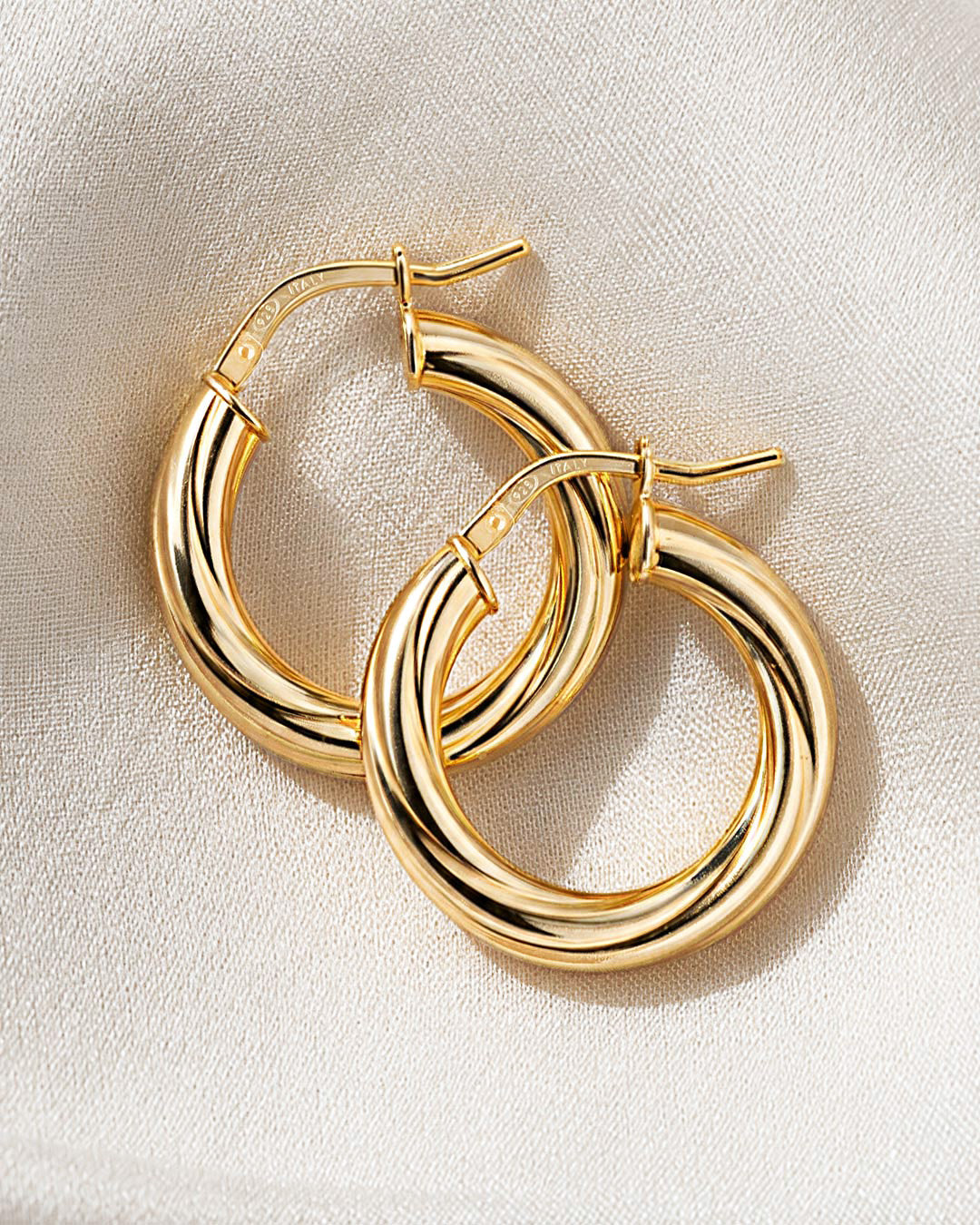 Pair of chunky gold tube hoop earrings with hinged closures, overlapping on cream woven fabric
