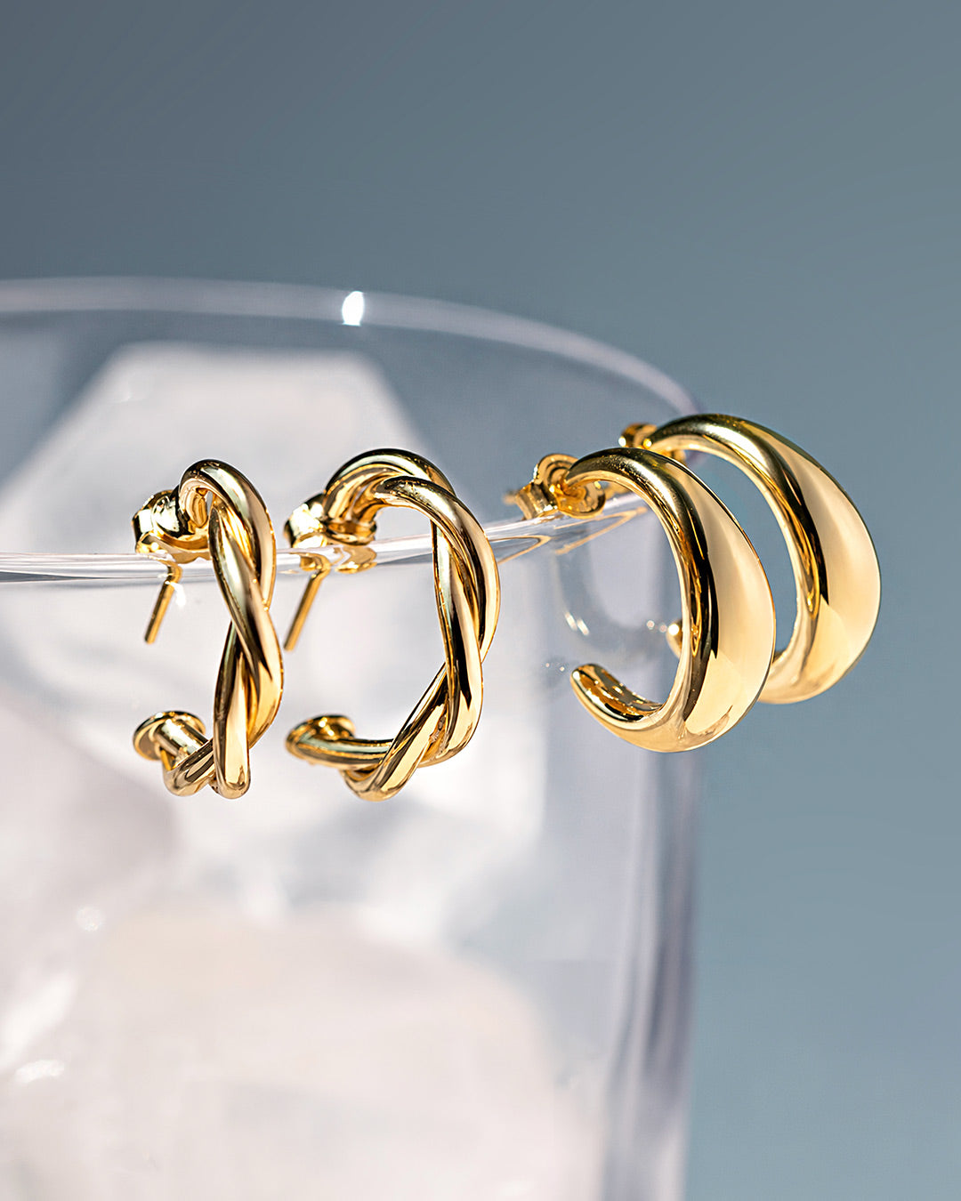 AMYO gold hoop earrings in twisted and chunky styles displayed on a glass, showcasing everyday hoops for effortless styling.