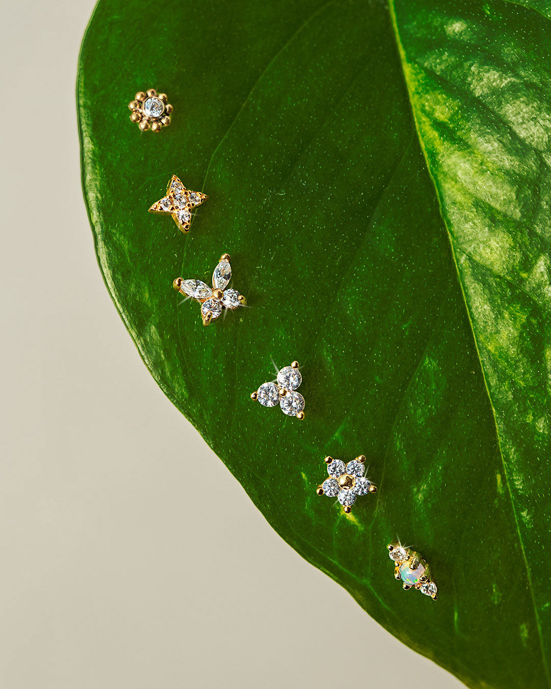 Dainty flat back earrings with crystal and gemstone designs displayed on a green leaf, featuring star, floral, and cluster styles. Linking to the AMYO flat back studs collection.