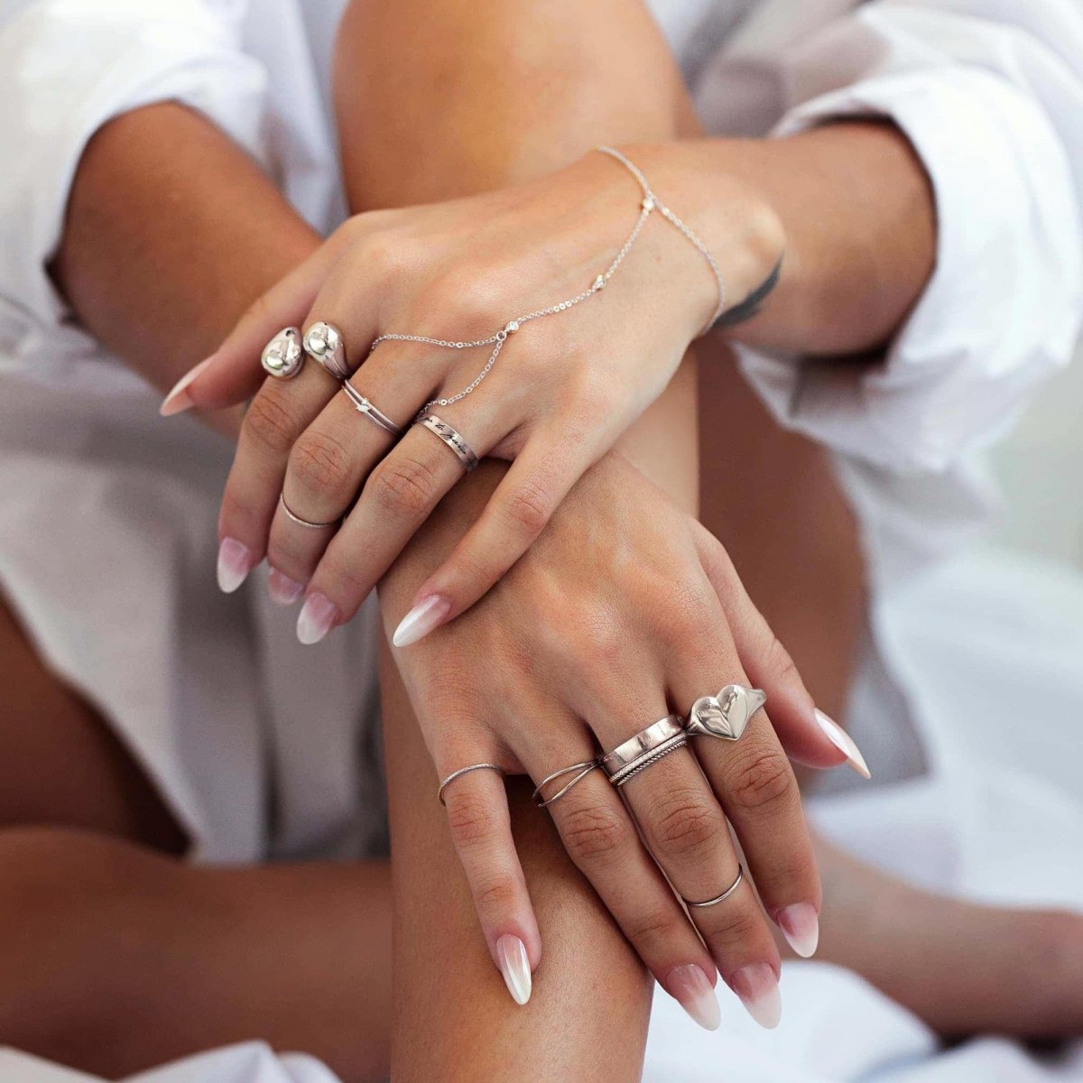 Woman wearing silver dainty crystal handchain, silver bold sculptural bypass ring, silver bold heart signet ring, silver tiny opal double band ring, silver cigar band ring, silver criss cross ring, silver pave eternity ring, and silver thin band and rope ring set