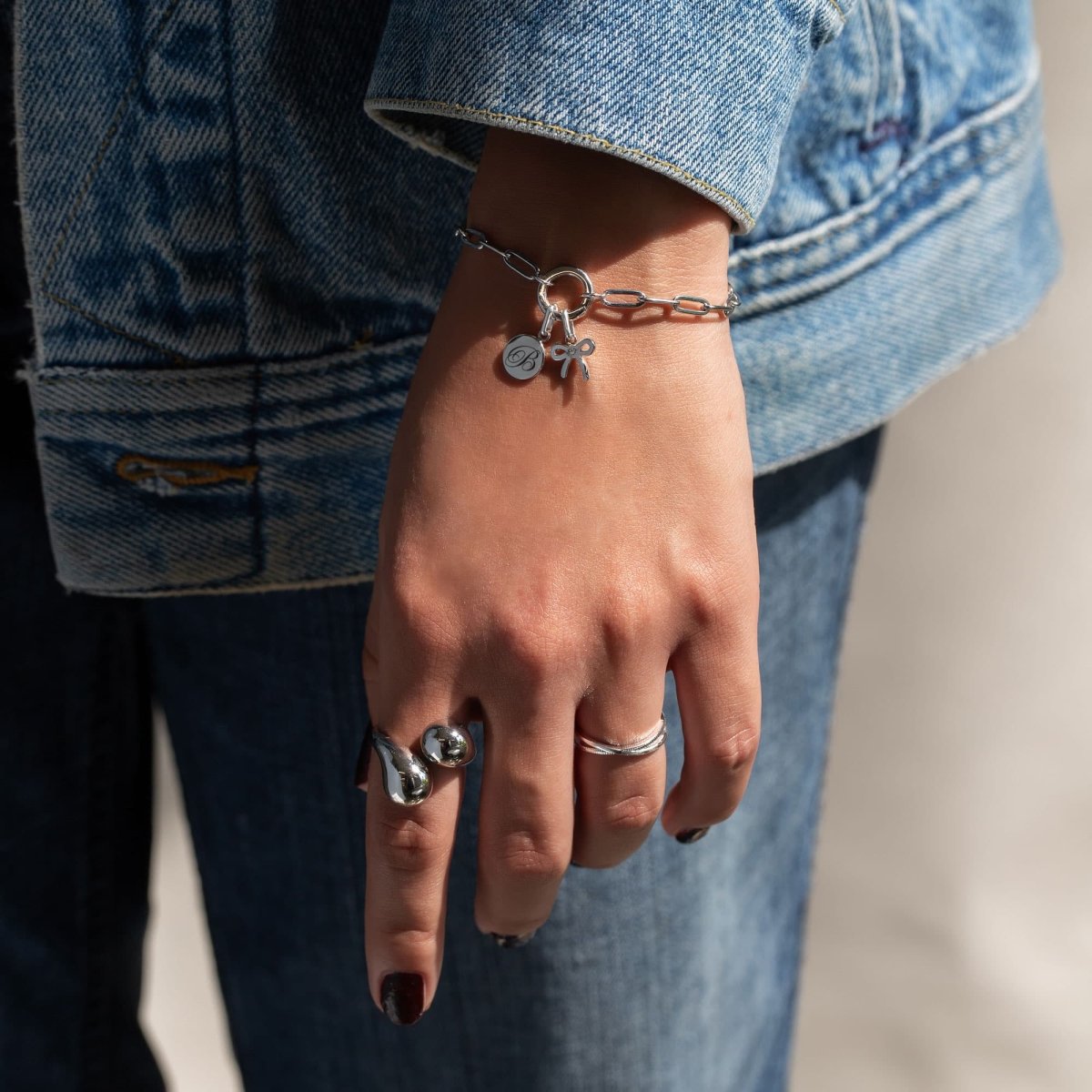 Sterling silver bow charm beside an initial disc charm on a chain bracelet, shown on a wrist