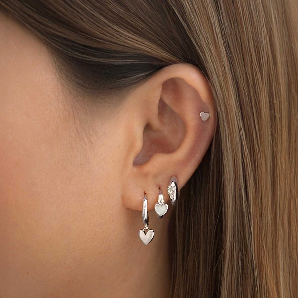 Close-up of an ear wearing heart-shaped huggie earrings and tiny heart stud earring from AMYO