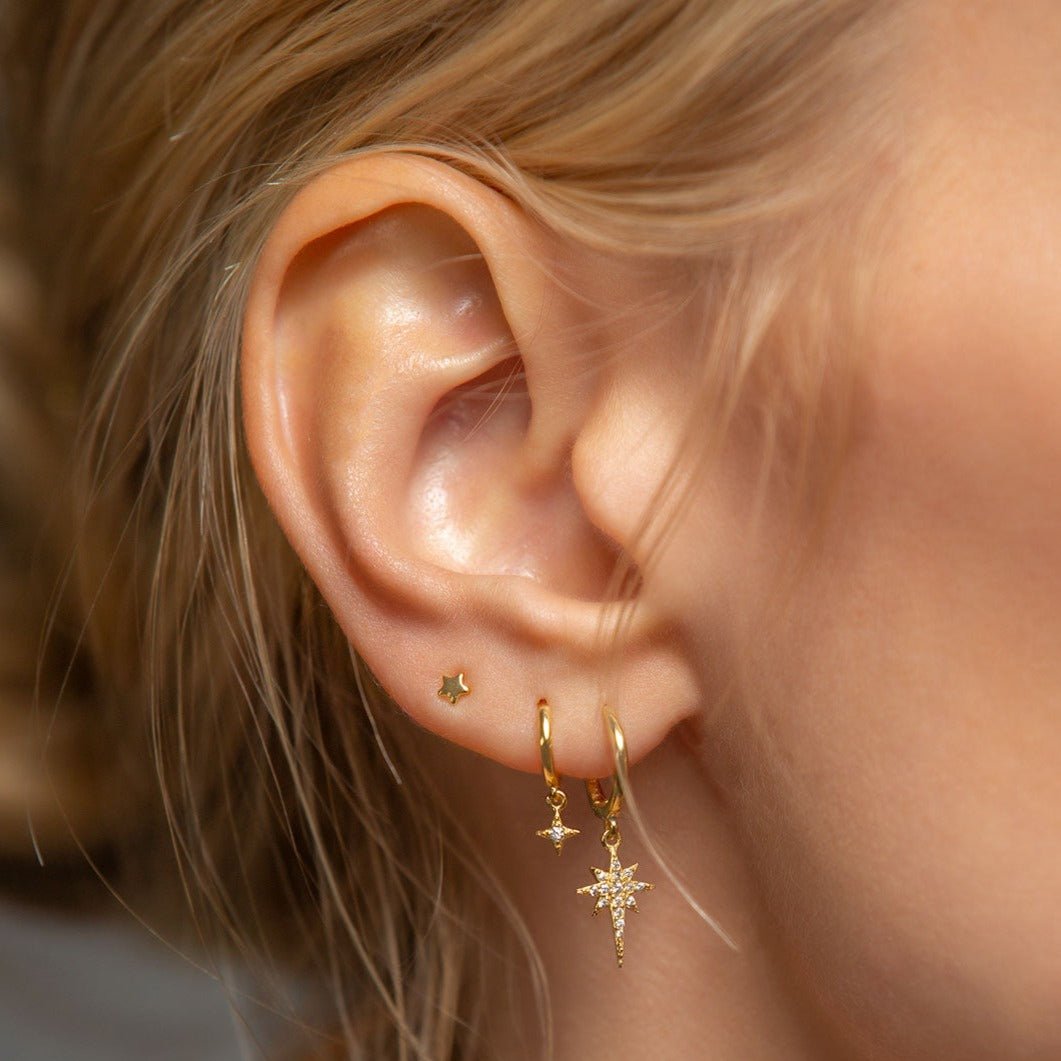 Close-up of woman wearing gold starburst huggie earring, gold star flare huggie earring, and gold tiny star stud