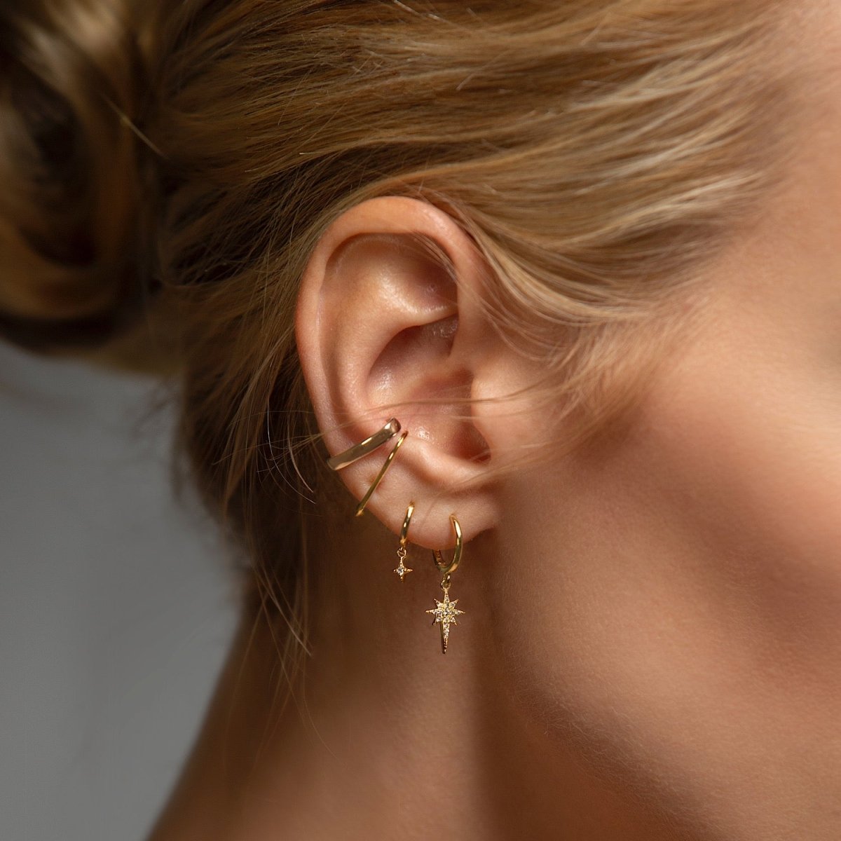 Close-up of woman wearing gold starburst huggie earrings, gold star flare huggie earrings, gold thin suspender earrings, and gold bold suspender ear cuff