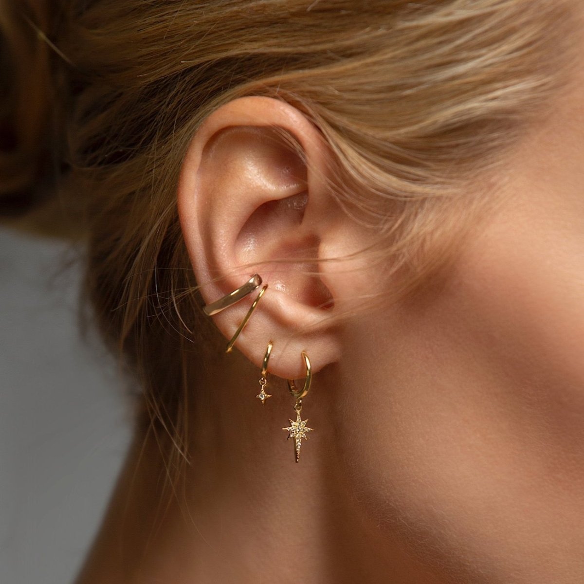 Close-up of woman wearing gold starburst huggie earring, gold star flare huggie earring, gold thin suspender earring, and gold bold suspender ear cuff