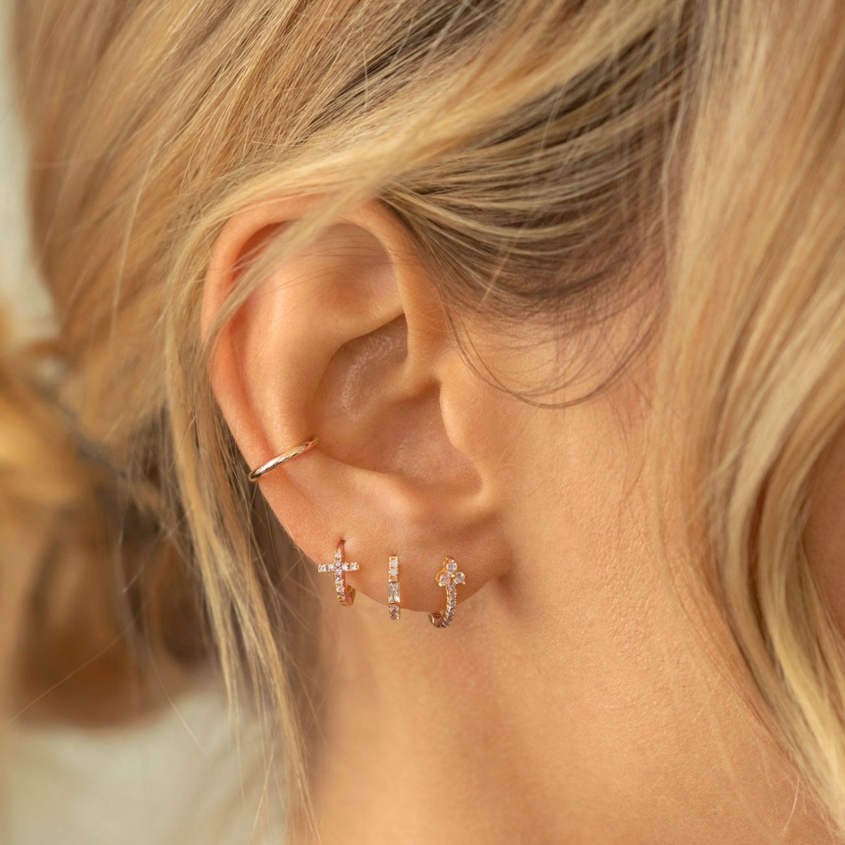 Close-up of woman wearing rose gold tiny cross huggie earrings, rose gold clover pave huggie earring, rose gold baguette huggie earrings, and rose gold eternity ear cuff