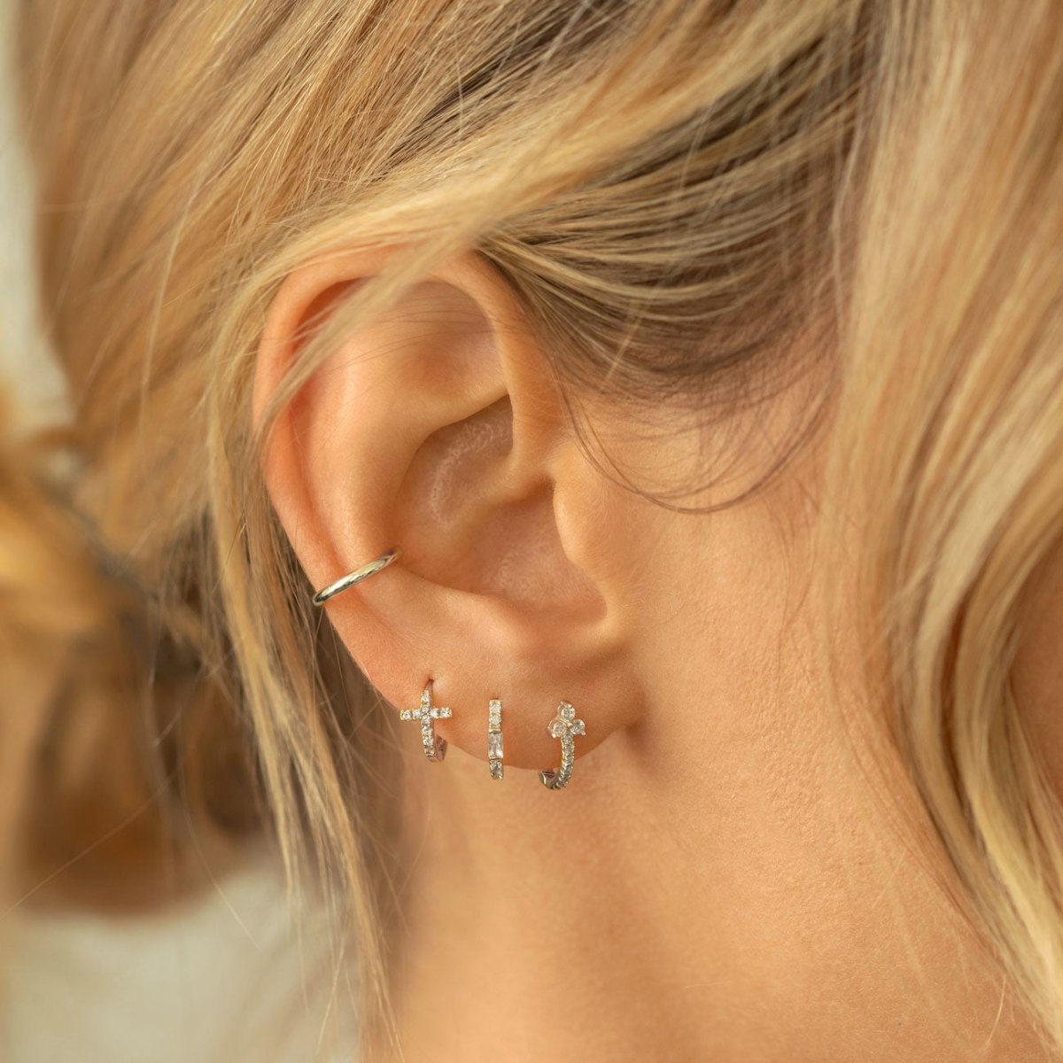 A close-up of woman's ear featuring sterling silver cross huggie earrings and conch cuff. 