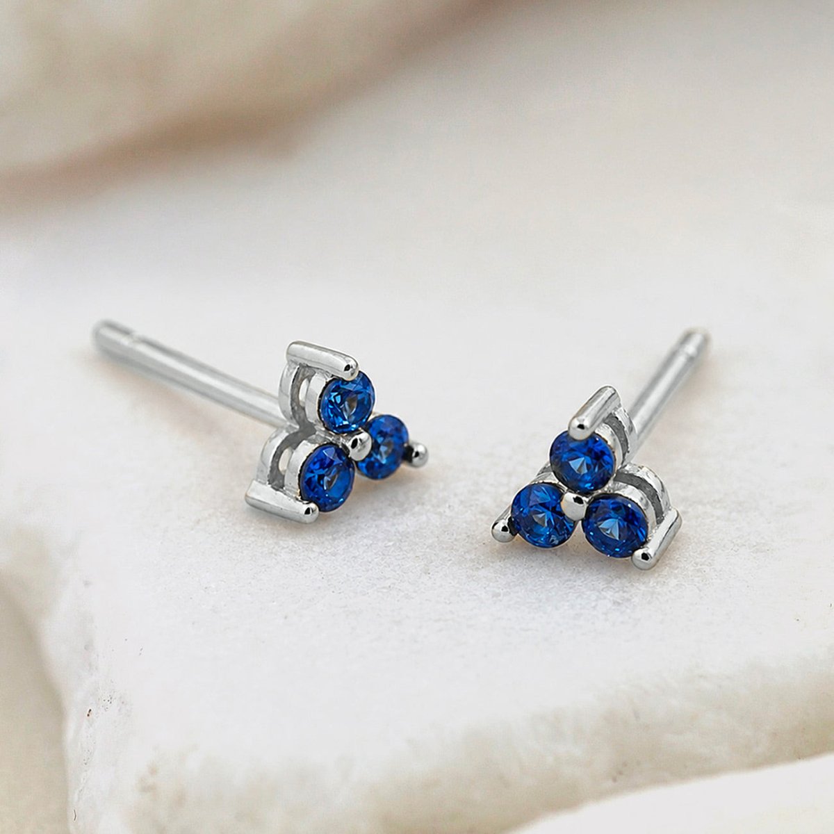 Pair of silver studs, each set with three round blue stones in a clover cluster, on a pale stone surface