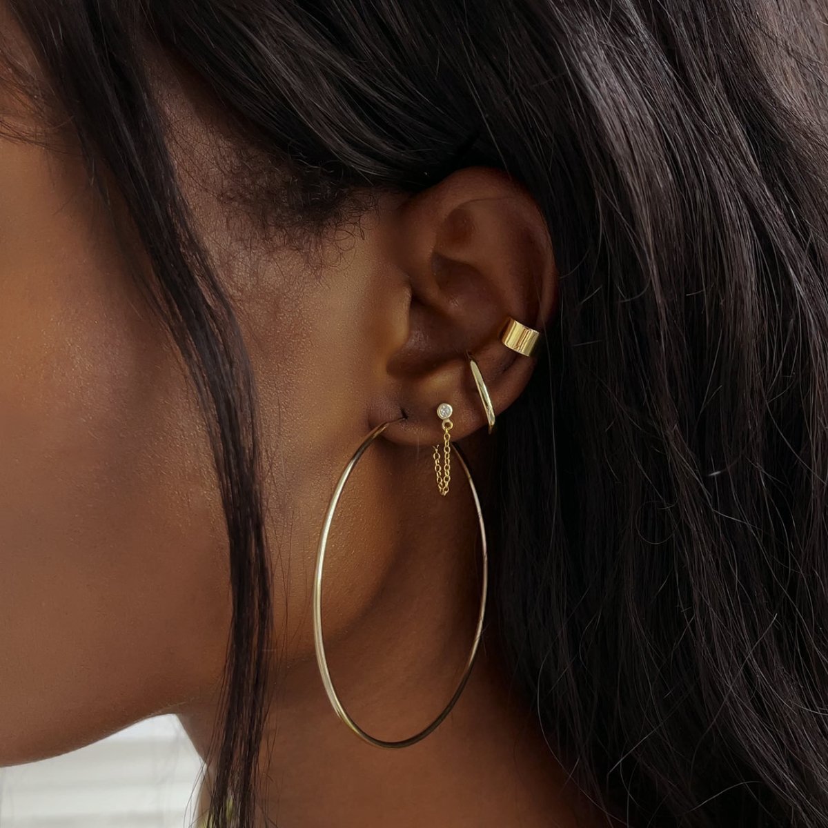 Close-up of woman wearing gold classic ear cuff, gold thin large hoops, gold suspender earrings, and gold bezel chain huggie earrings