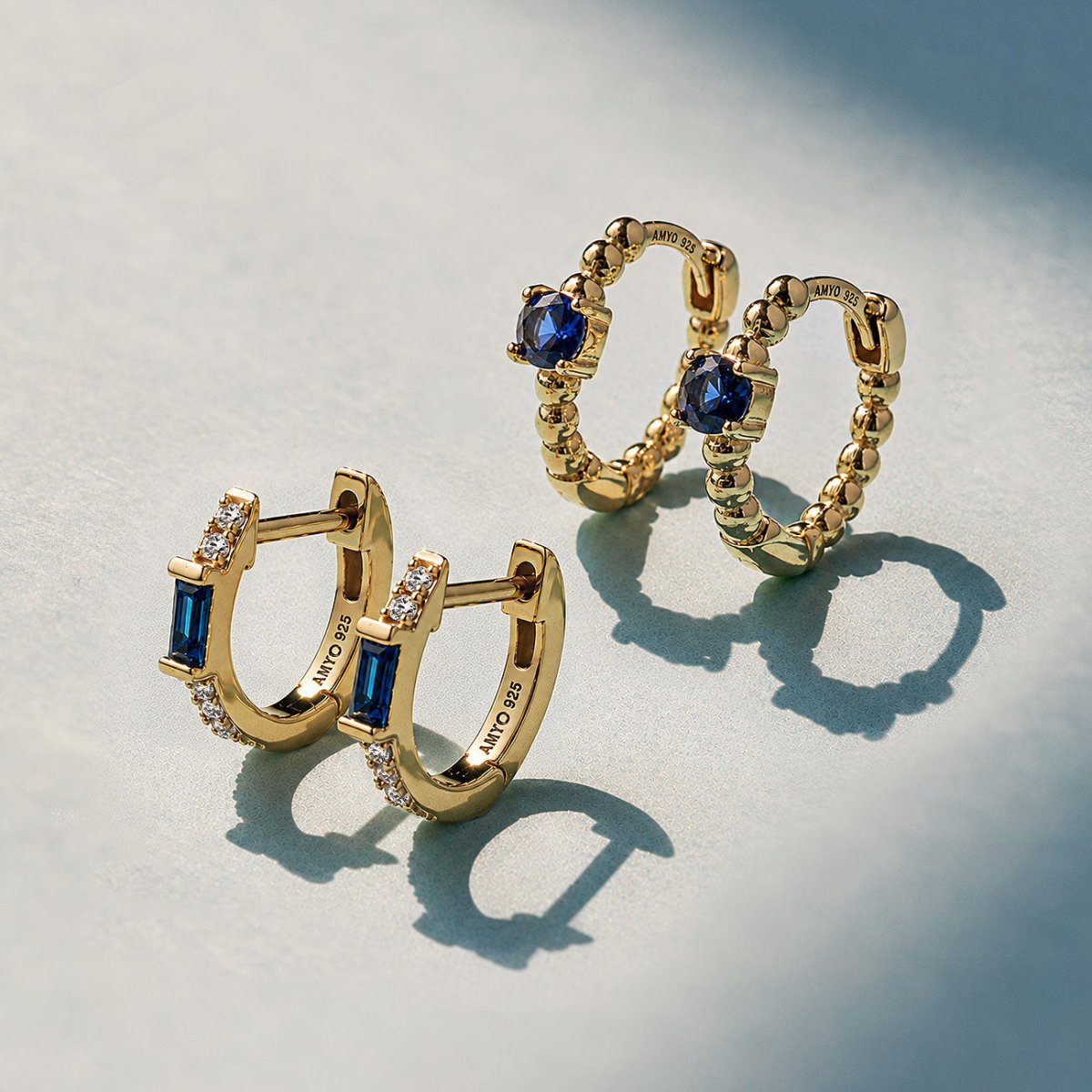 Two pairs of gold vermeil huggie hoops with blue baguette and round stones on a pale surface