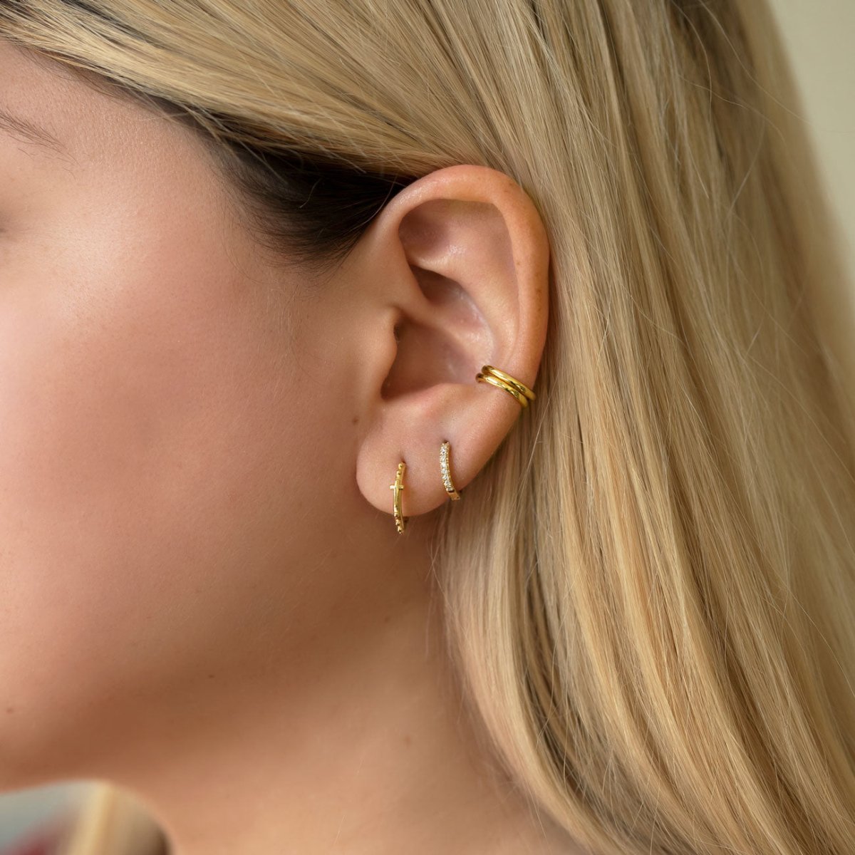 Close-up of woman wearing gold Eternity Ear cuff, cross bead mini hoops, thin pave huggie hoops
