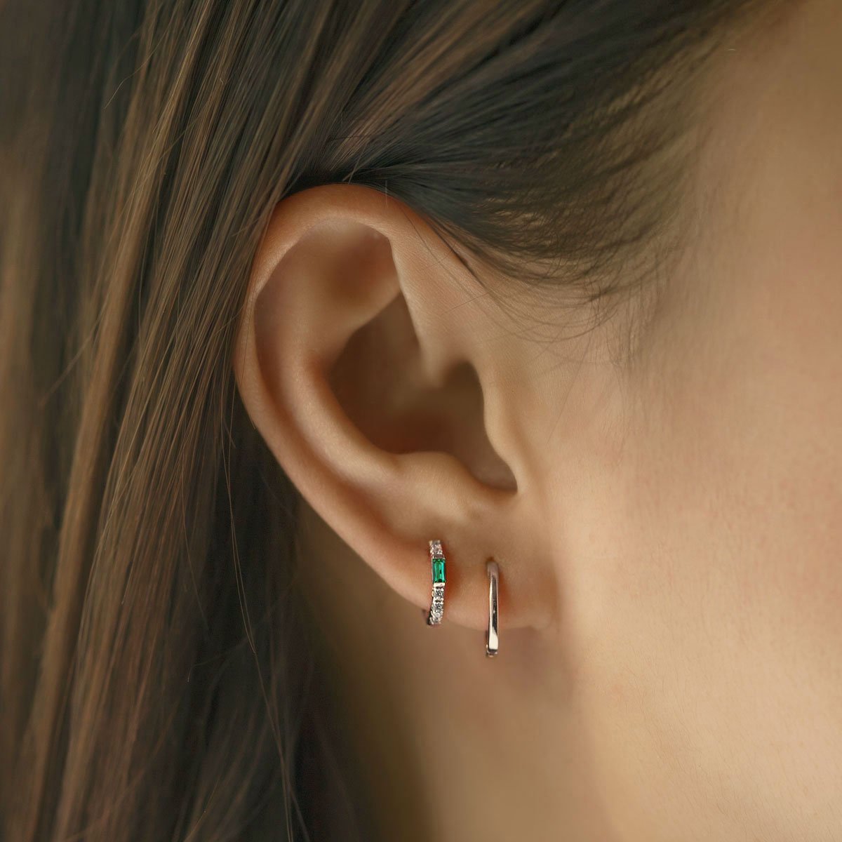 Close-up of woman's ear featuring Sterling Silver Gemstone Baguette Huggie Earrings Emerald and silver hoops. 