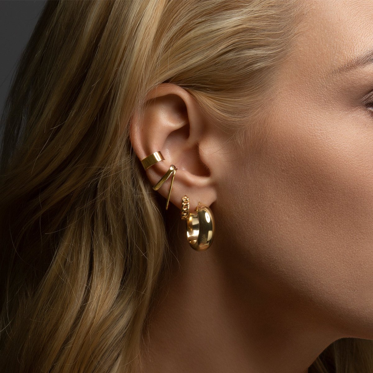 Close-up of woman wearing gold chunky hoops, gold mini chain link hoops, gold bold suspender ear cuff, gold thin suspender earrings, and gold classic ear cuff