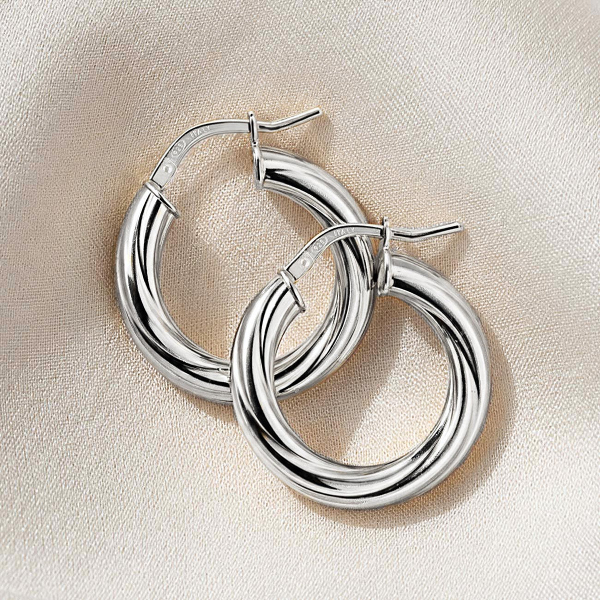 Pair of thick twisted silver hoop earrings with hinged latch closures, overlapping on cream fabric