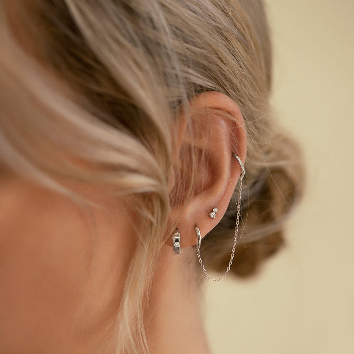 Close-up of woman's ear featuring 14k white gold thick milgrain diamond huggie, sterling silver tiny two diamond single stud, 14k white gold single mini huggie, and 14k white gold cartilage pave hoop earring