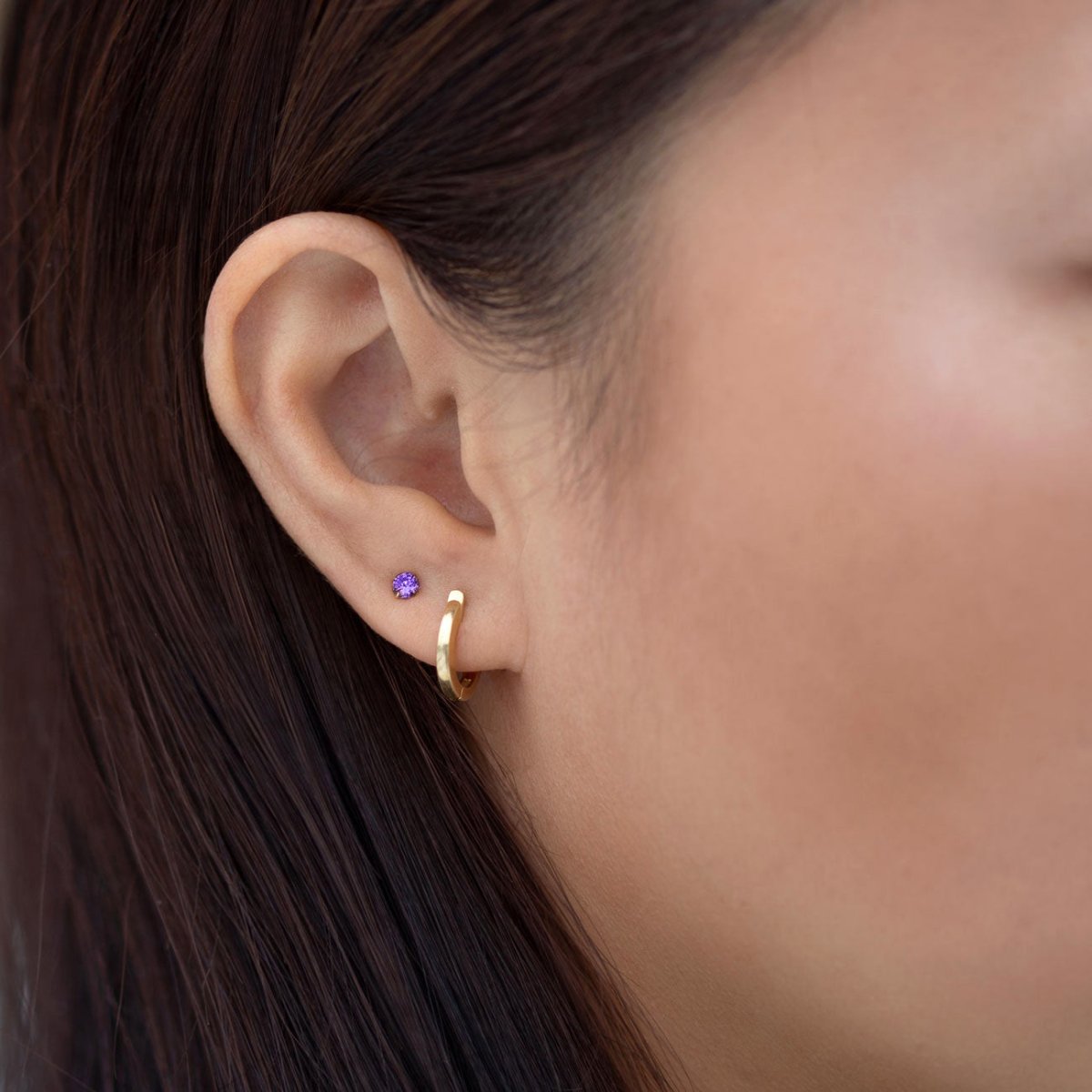 Close-up of woman's ear featuring 14K Gold Birthstone Tiny Studs Amethyst paired with huggie earring 