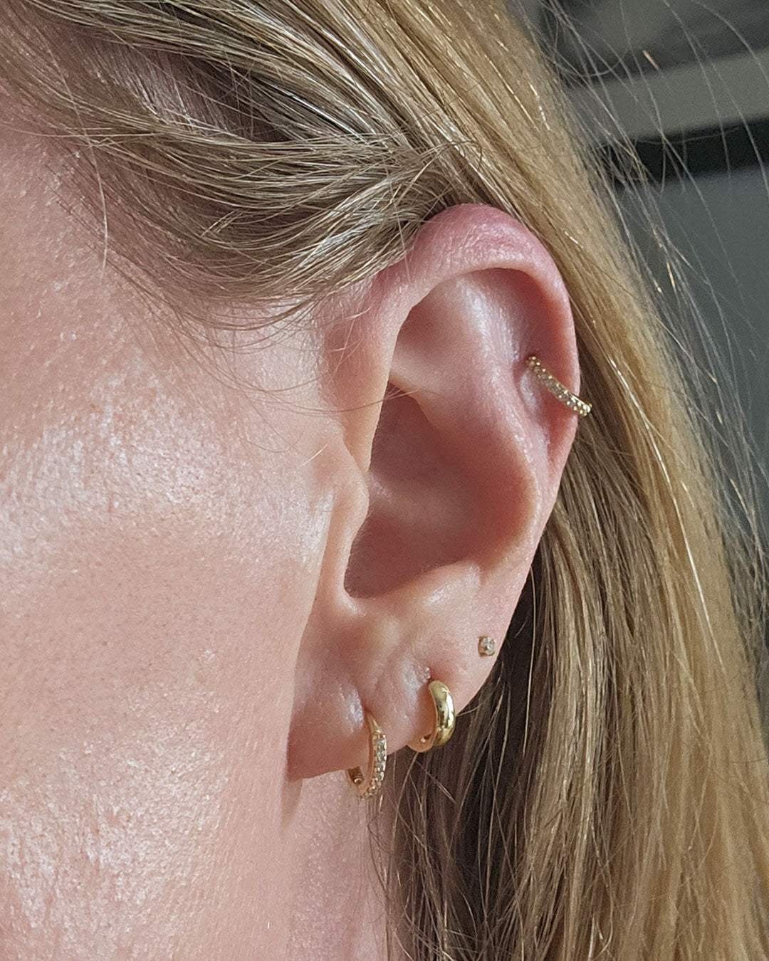 Close-up of an ear wearing a gold crystal-set helix hoop, a small stud and two gold lobe huggie hoops