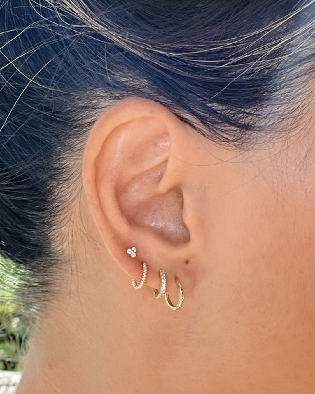 Close-up of an ear wearing a three-stone cluster stud with a twisted gold huggie, a stone-set huggie and a plain hoop