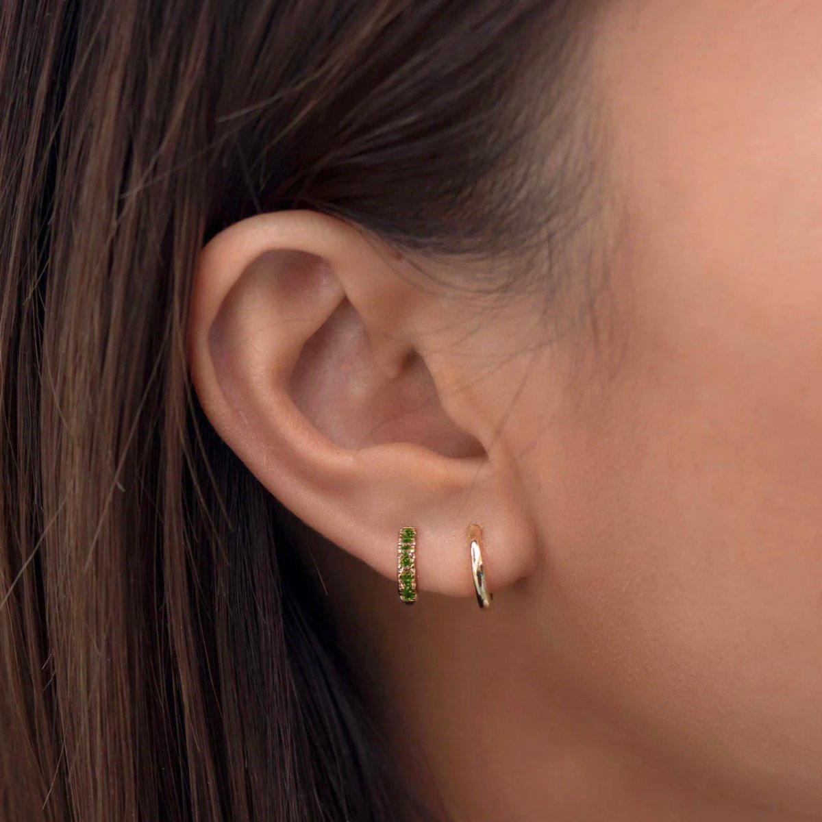 Close-up of woman's ear featuring 14K gold pave huggie emerald crystal earring. 