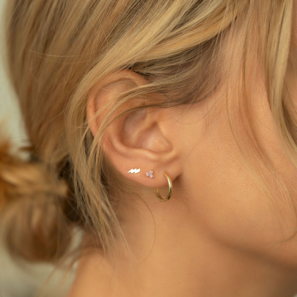 Close up of woman's ear featuring 14K Gold Gemstone Clover Studs Pink CZ Crystals, lightning bolt stud, and gold hoop earring 