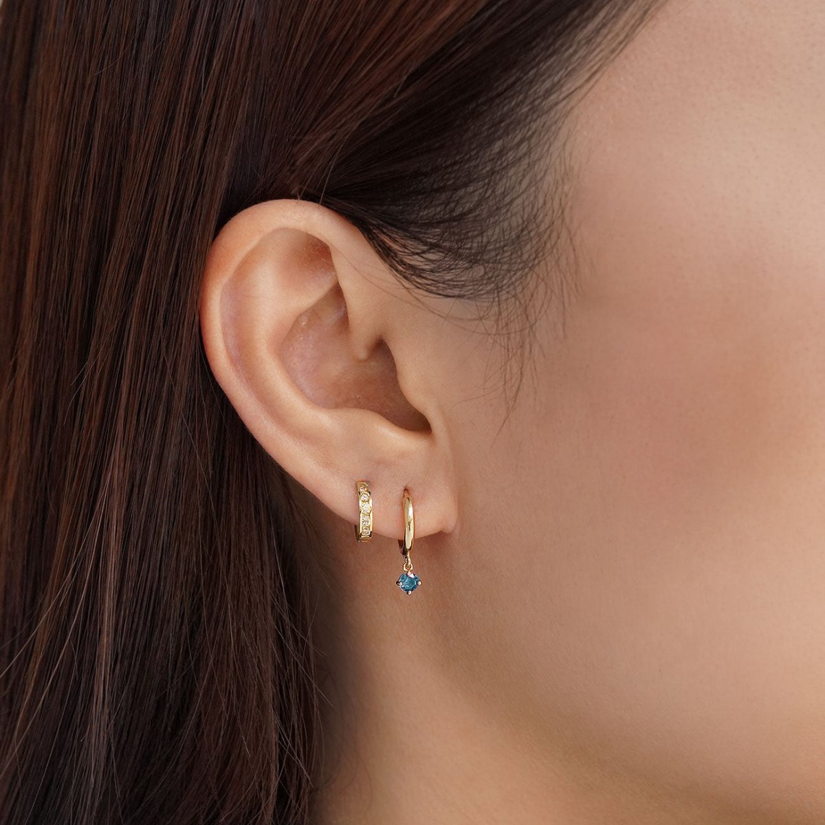 Close-up of woman's ear featuring 14K Gold Gemstone Dangle Huggies Blue Zircona CZ Crystal paired with huggie earring. 