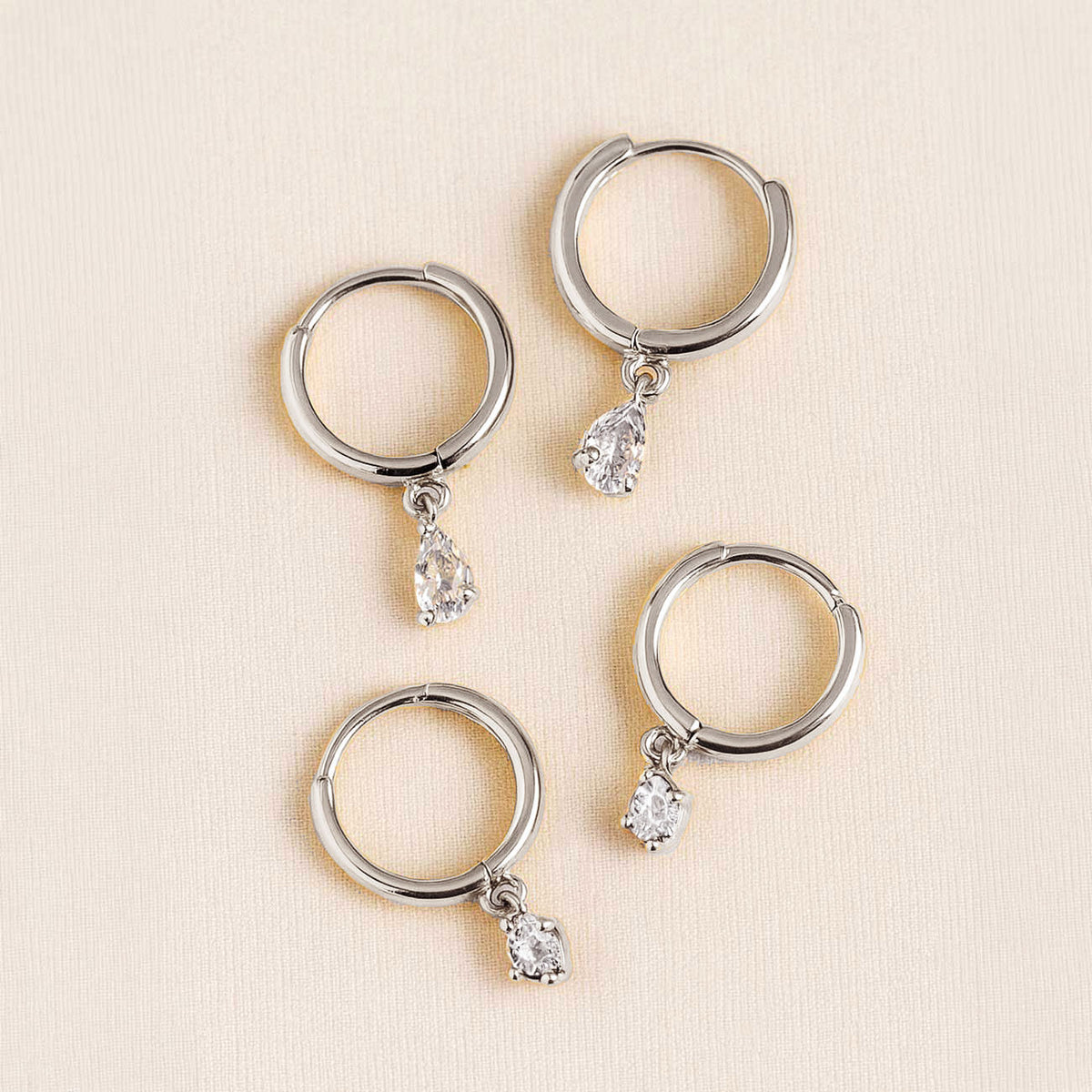 Four 14K white gold huggie hoops with pear-shaped and round moissanite drops on a cream surface