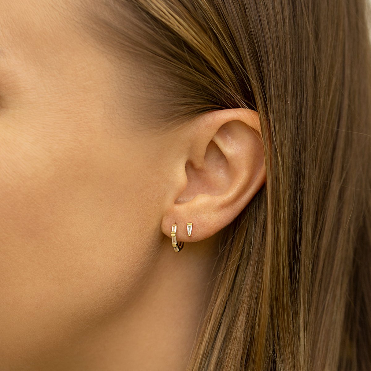 Close-up on woman's ear featuring Amyo Jewelry's 14K Solid Gold Tri Baguette Huggie Hoops Moissanite Crystals paired with stud earring