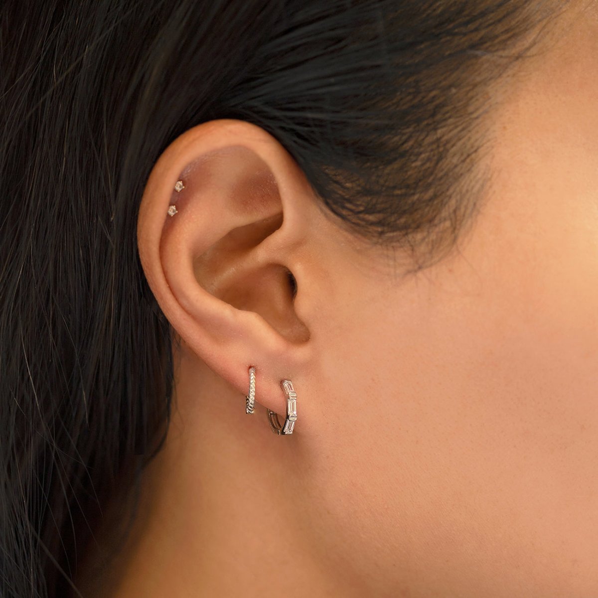 Close-up of woman's ear featuring White Gold Texture Rope Huggie Hoop Earrings and 14K White Gold Tri Baguette Huggie Hoops