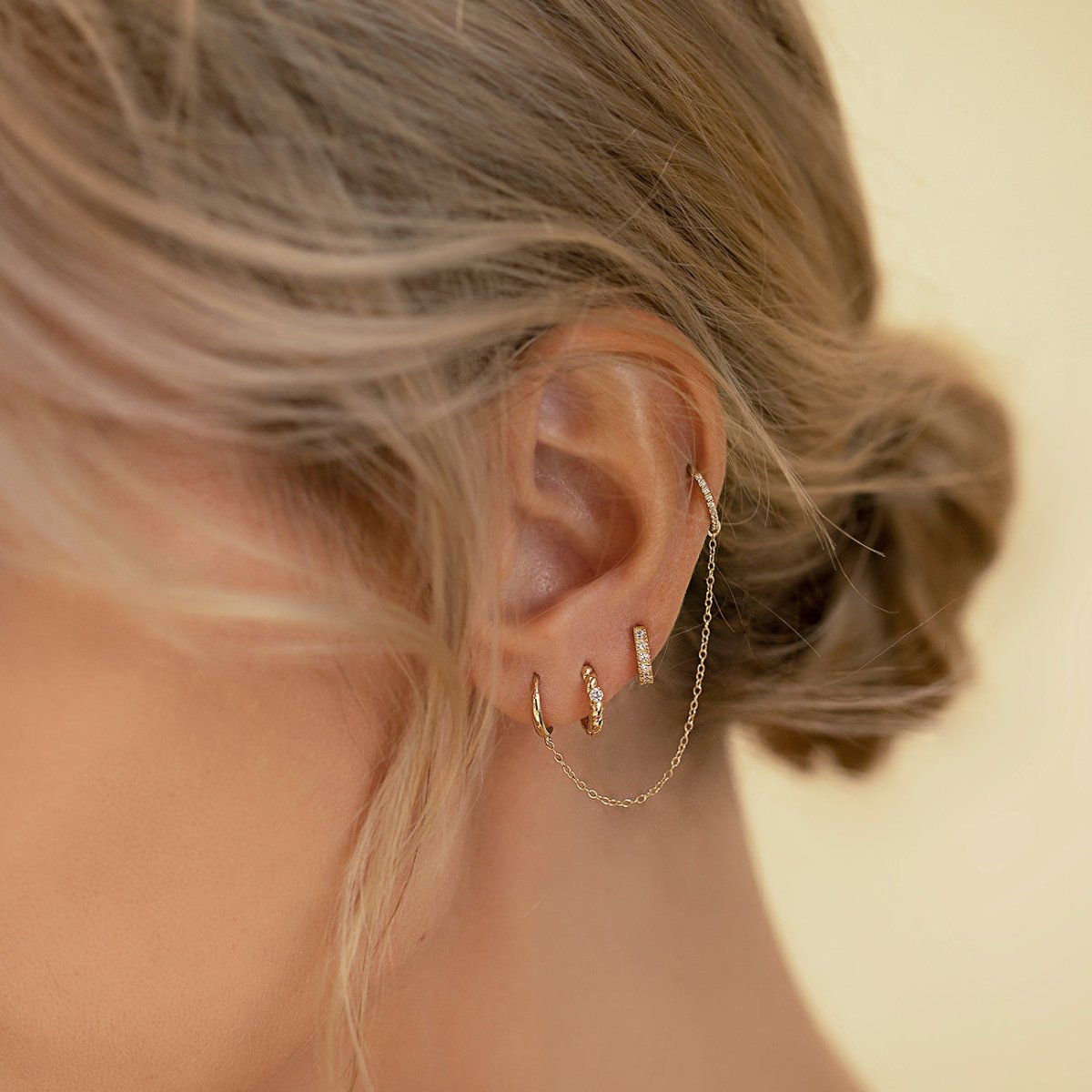 Close-up of woman's ear featuring 14K gold moissanite crystal pave huggie earrings and a chain set of 14K gold earrings. 